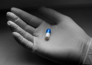 白い医療用手袋をした手が、青と白のカプセル状の薬を静かに持っている様子。清潔感のある背景に、薬品管理や医療現場の緊張感が感じられる構図。