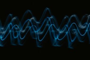 黒い背景に青い光の波形が重なりながら浮かび上がる様子。周波数、電磁波、脳波、5G、音響技術、周波数療法、または振動と意識の関係性を象徴。