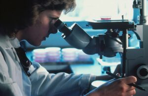 顕微鏡をのぞいて研究を続ける白衣姿の科学者の横顔。手元には操作用の機器があり、真剣な眼差しで観察している様子。