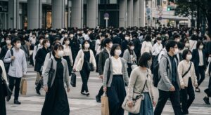 日本の都市街頭でマスクをした群衆が無言で歩く様子。整然と同じ方向に進む姿が、“従順さ”と“同調圧力”の象徴として描かれ、支配層にとっての“管理しやすい国”であることを暗示。統治実験に最適な社会性を示す視覚表現。